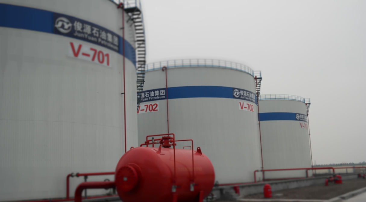 Industrial facility with large Cyclopentane storage tanks labeled V-701, V-702, and V-703, belonging to Junyuan Petroleum Group. Each tank features a blue identification band and access ladders. A red horizontal pressure vessel with piping and valves is visible in the foreground, indicating safety or fire suppression infrastructure. The setting is overcast, with a concrete base and organized layout.
