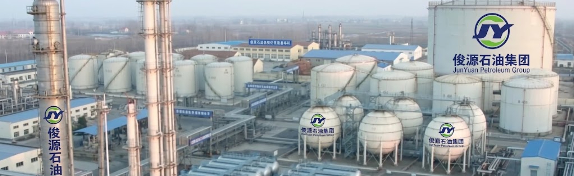 An aerial view of the JunYuan Petroleum Group industrial chemical processing facility featuring large storage tanks, spherical gas containers, tall distillation towers, and complex pipeline infrastructure under a clear sky.