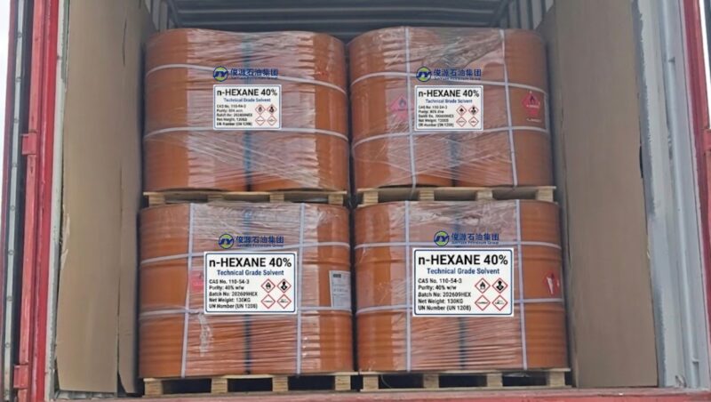 Four palletized stacks of orange industrial steel drums containing 40% n-hexane (CAS 110-54-3) with GHS hazard labels, loaded inside a cargo shipping container.